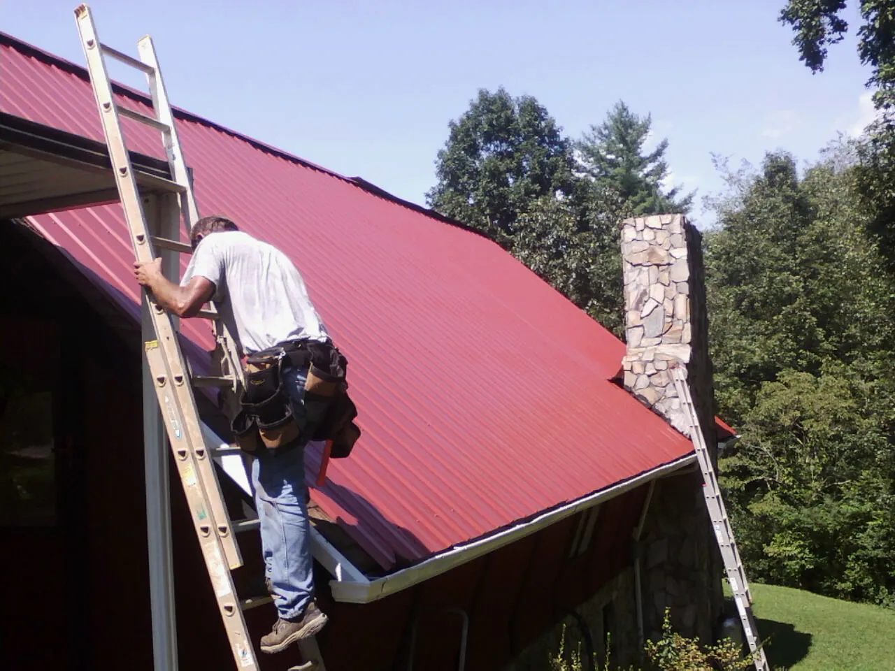 Roofer on a ladder beside a red metal roof and stone chimney — Soffit & Fascia Repair by Roofing Brothers in Grand Island